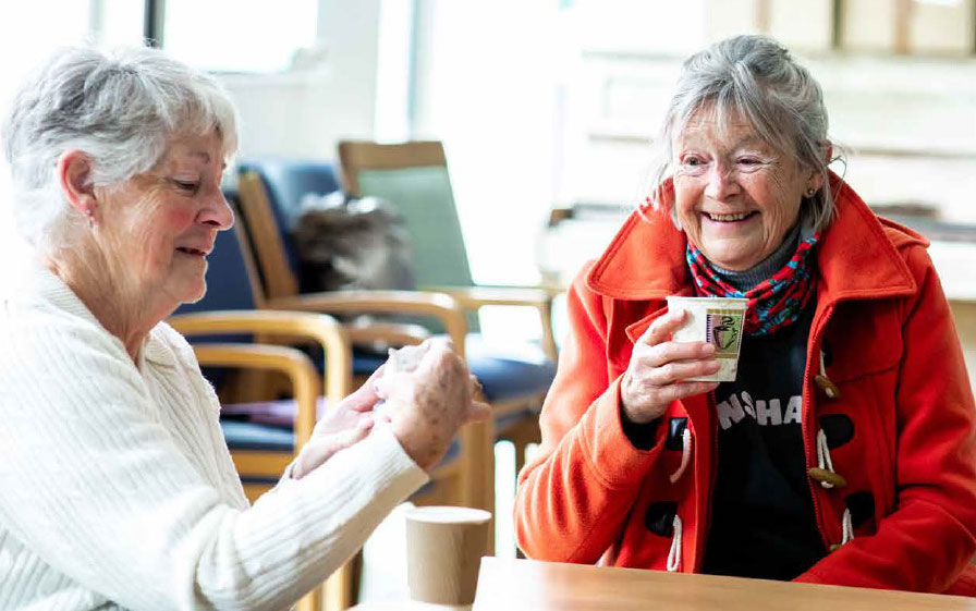 Whit-case-study Two women are sitting at the corner of a table, each with a hot drink. They are smiling.