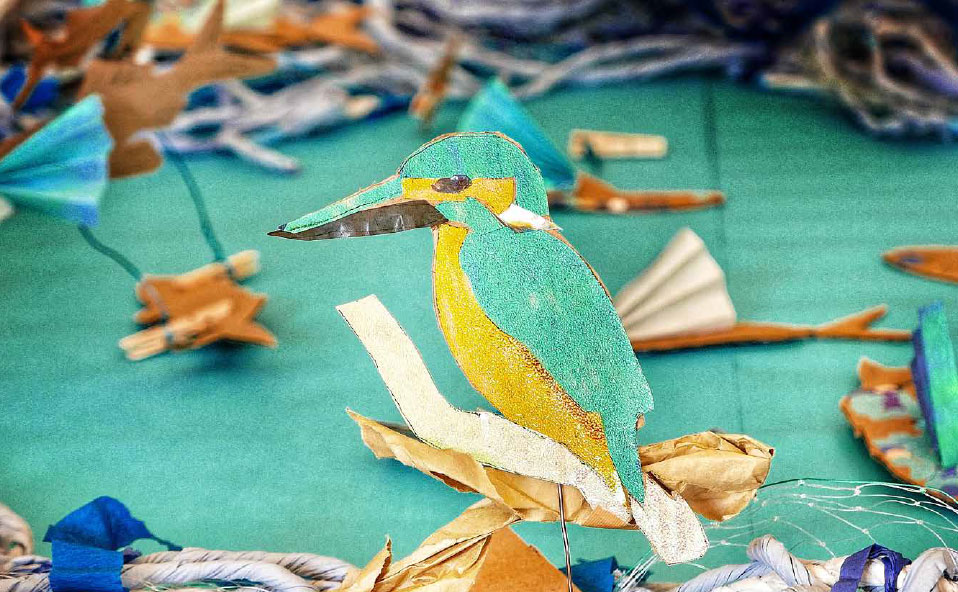 impact-footer a sculpture of a kingfisher with blue, orange and white colouring is in front of a teal fabric